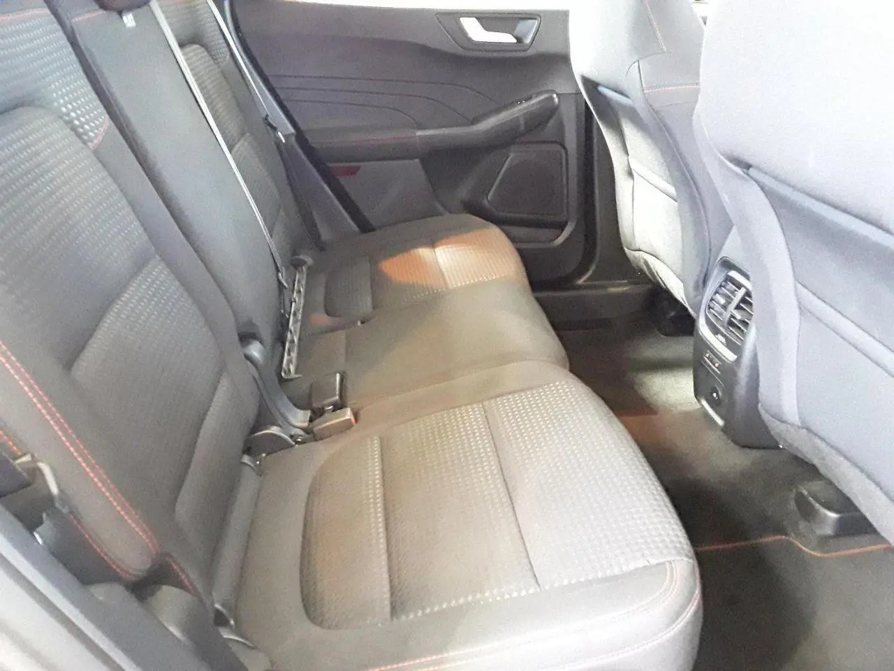 Vue intérieure arrière côté gauche du Ford Kuga 2024, sièges tissu gris avec surpiqûres rouges et aérateurs centraux.
