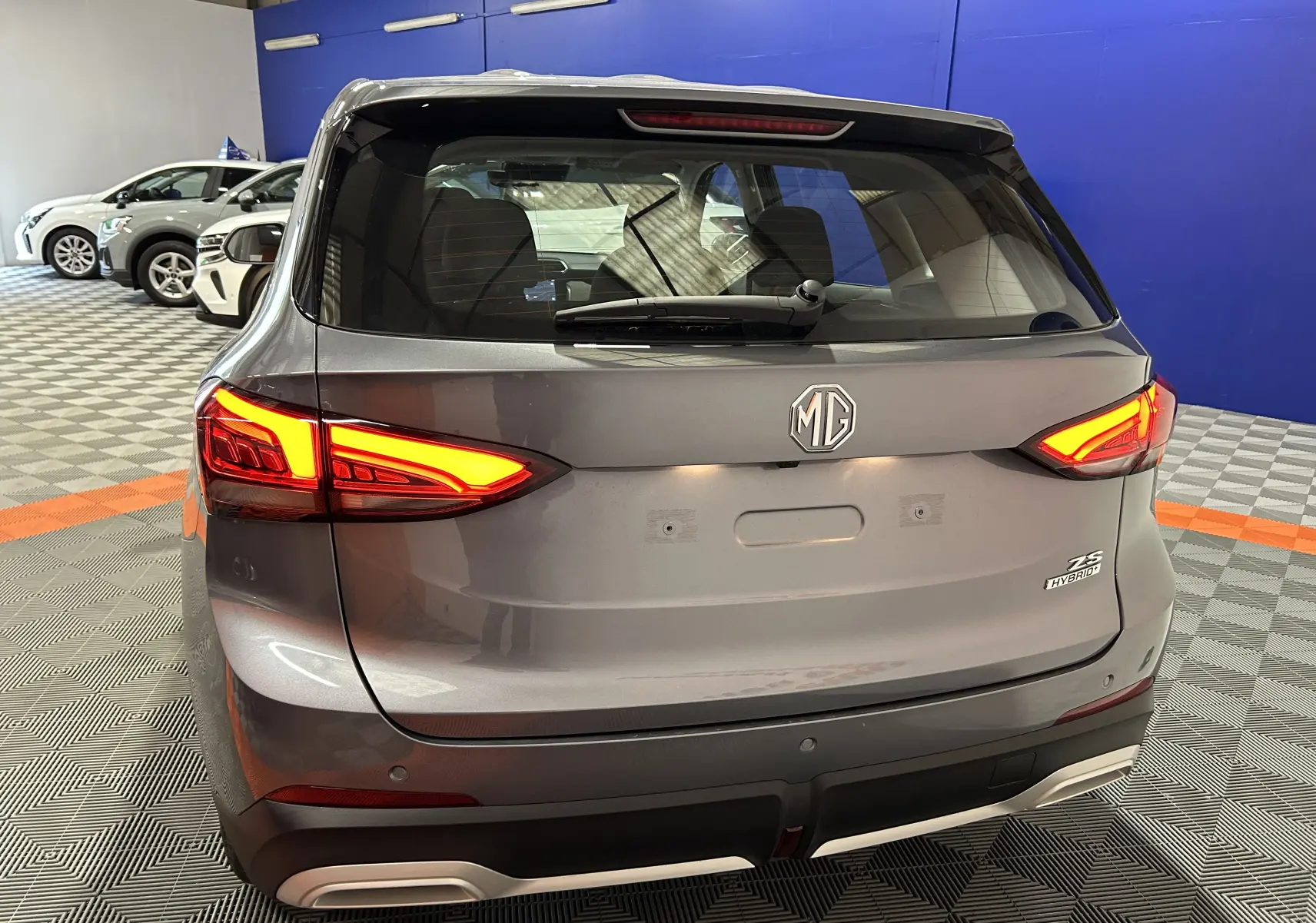 Vue arrière du MG ZS Hybrid+ 197 en gris Hampstead, avec feux allumés et logo MG bien visible.