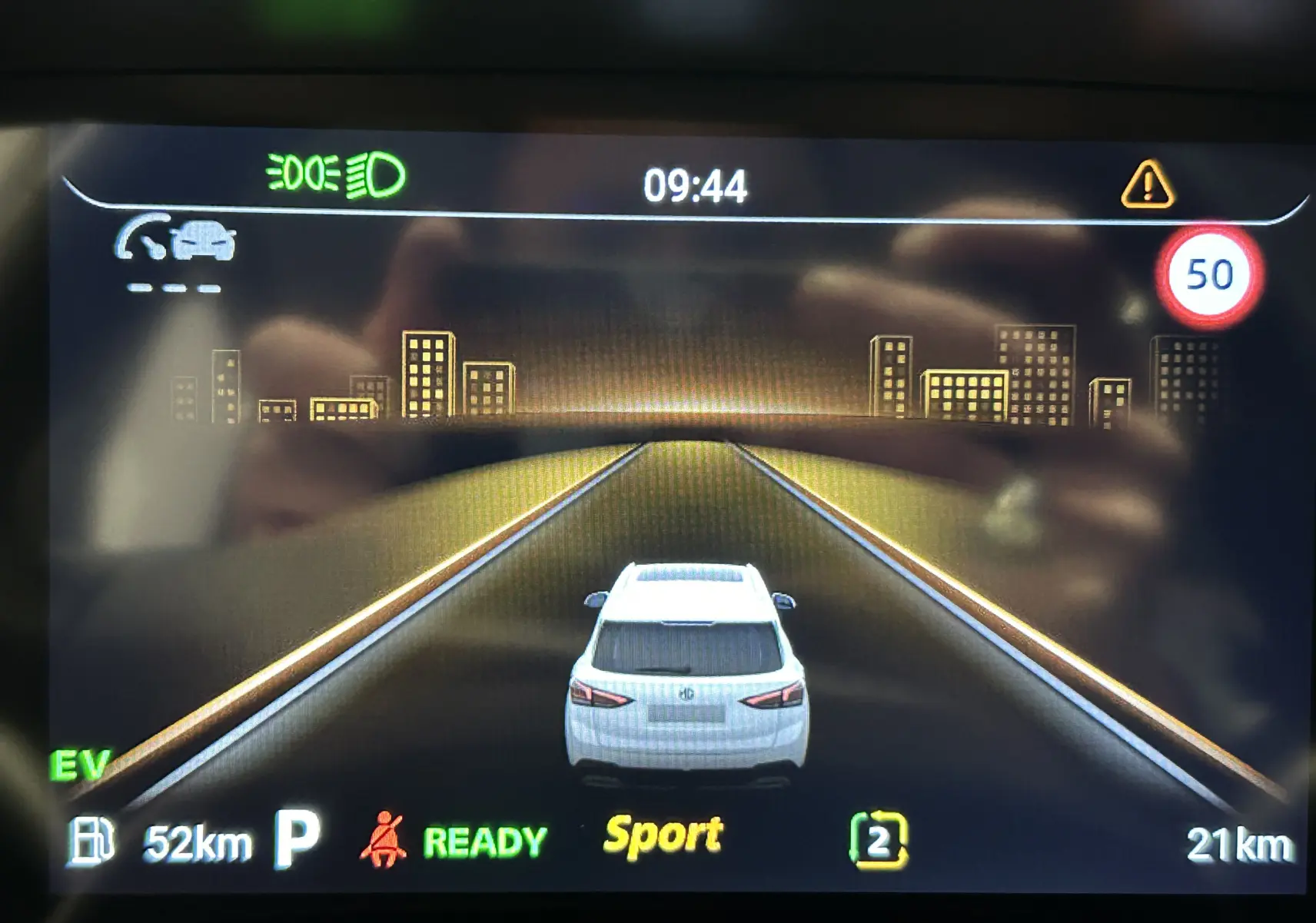 Affichage numérique montrant la voiture blanche MG ZS Hybrid+ vue de l'arrière sur une route virtuelle avec limite à 50 km/h.