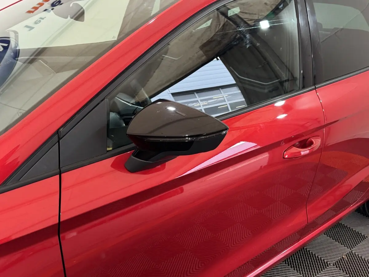 Vue rapprochée du côté gauche d’une SEAT Ibiza 1.0 TSI 110 FR rouge, mettant en valeur le rétroviseur noir et la poignée de porte.
