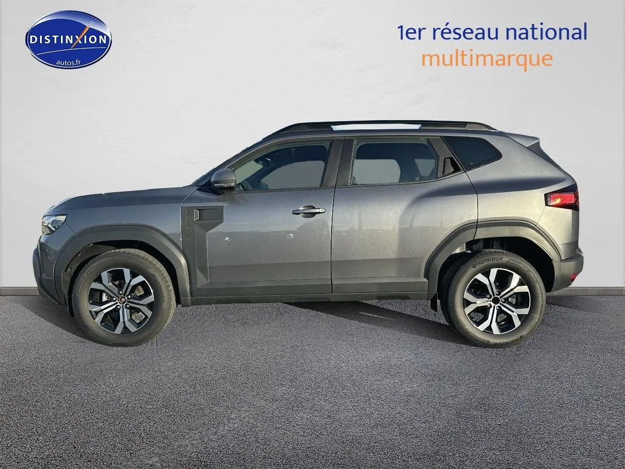 Vue de profil côté gauche d'un Dacia Duster gris schiste 2025 avec jantes alu et barres de toit noires.