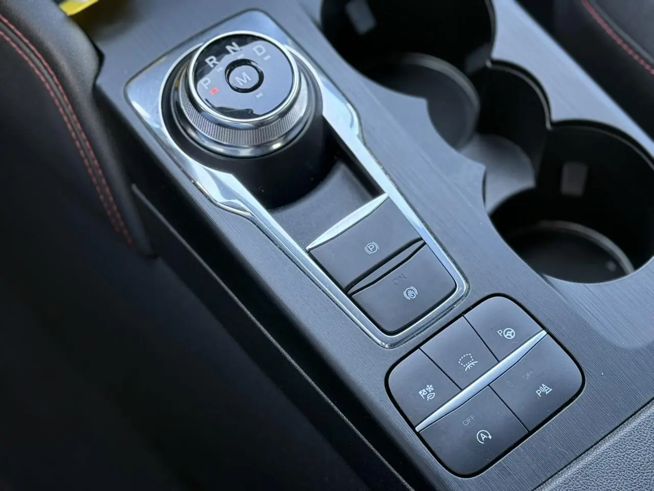 Gros plan sur la console centrale noire du Ford Kuga 2024 avec sélecteur rotatif de vitesse et boutons d’assistance au stationnement.