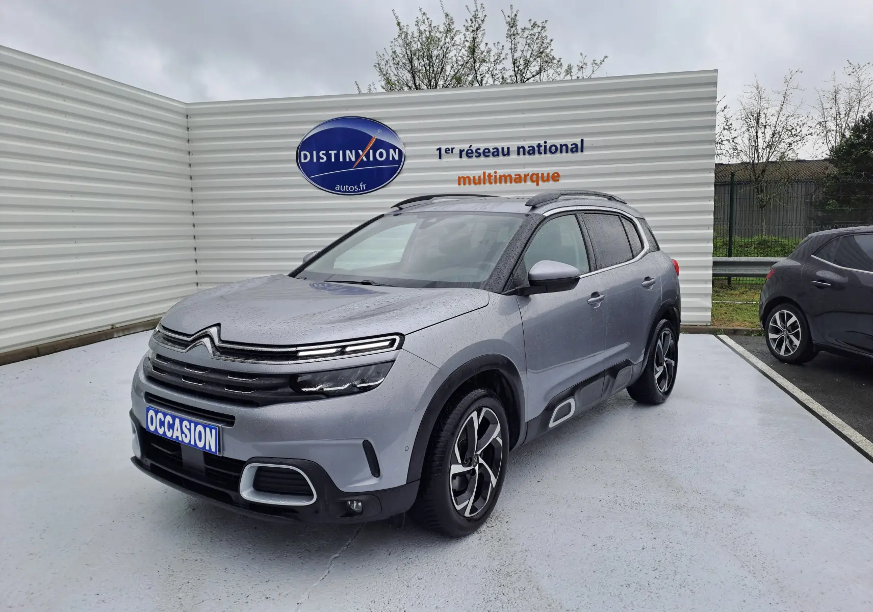 SUV Citroën C5 Aircross gris Artense vu en 3/4 avant droit sous la pluie avec jantes bi-ton et barres de toit.