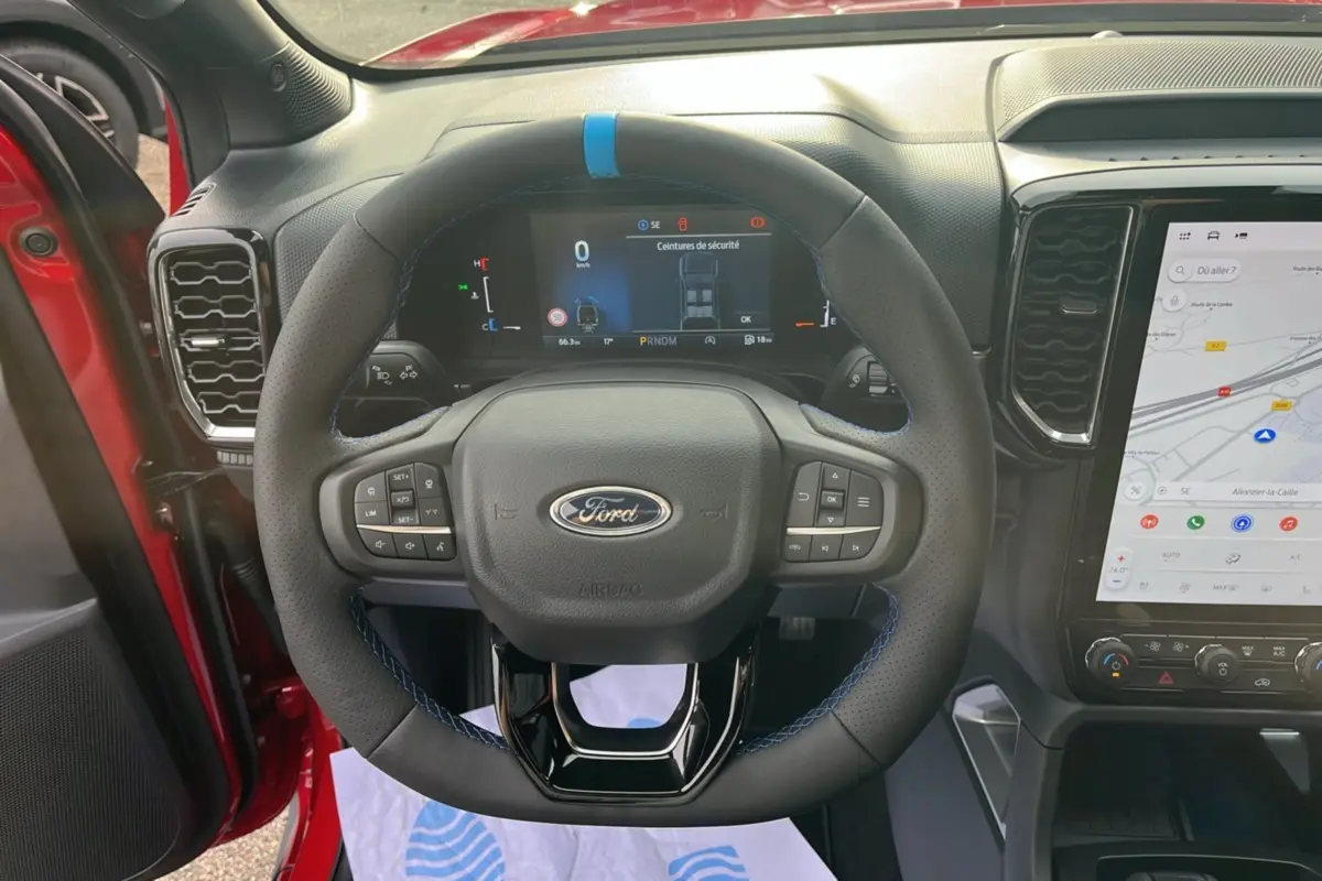 Vue rapprochée du volant cuir noir avec surpiqûres bleues du Ford Ranger 3.0 V6 Lucid Red, tableau de bord numérique allumé.