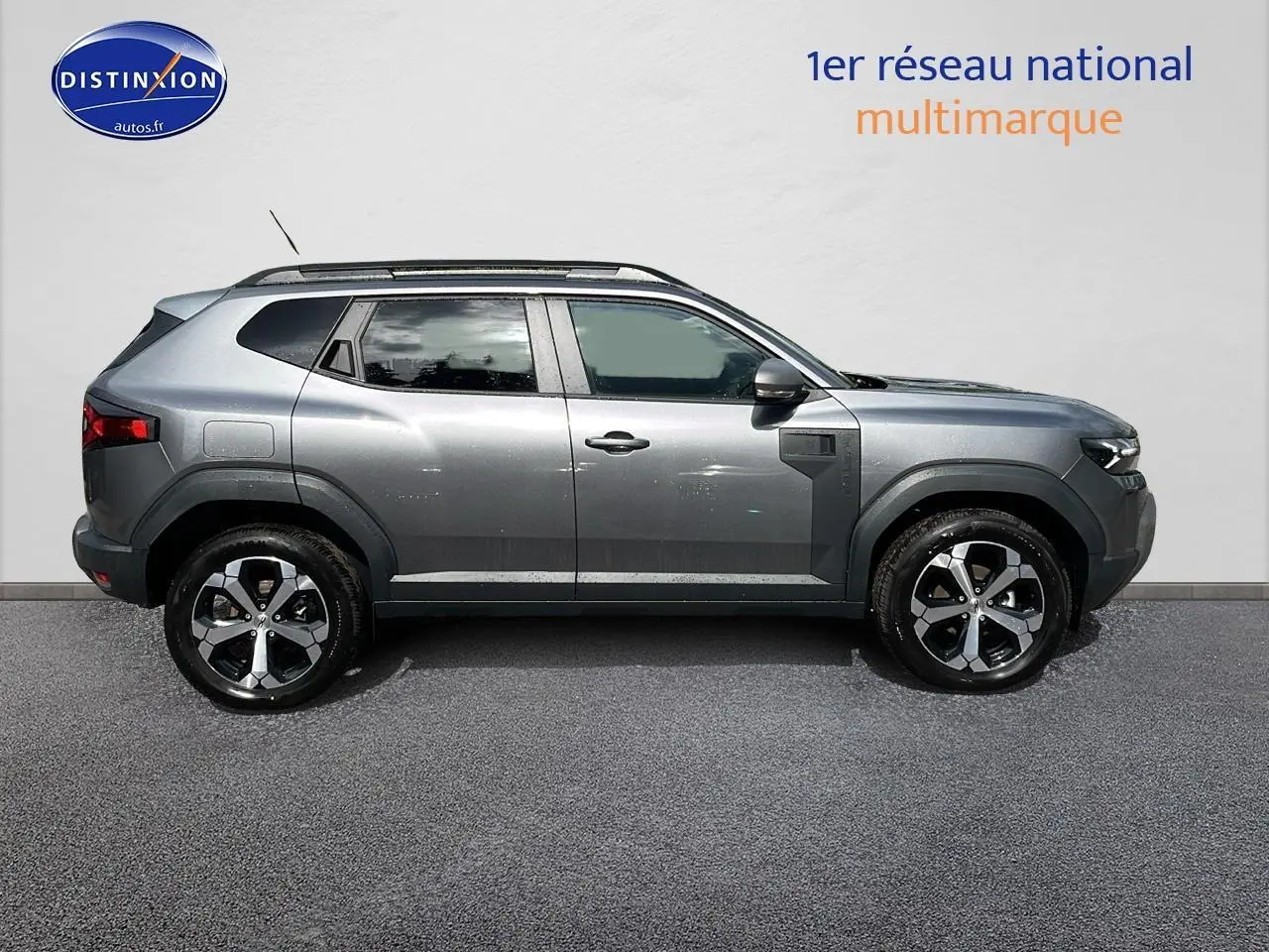 Profil droit du Dacia Duster gris schiste 2025 avec jantes alu et barres de toit visibles sur fond neutre.