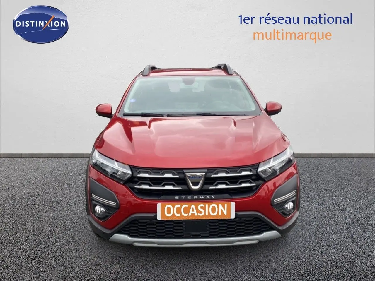Vue frontale d'une Dacia Sandero Stepway rouge fusion métal avec calandre noire et badge central, en arrière-plan un mur blanc.