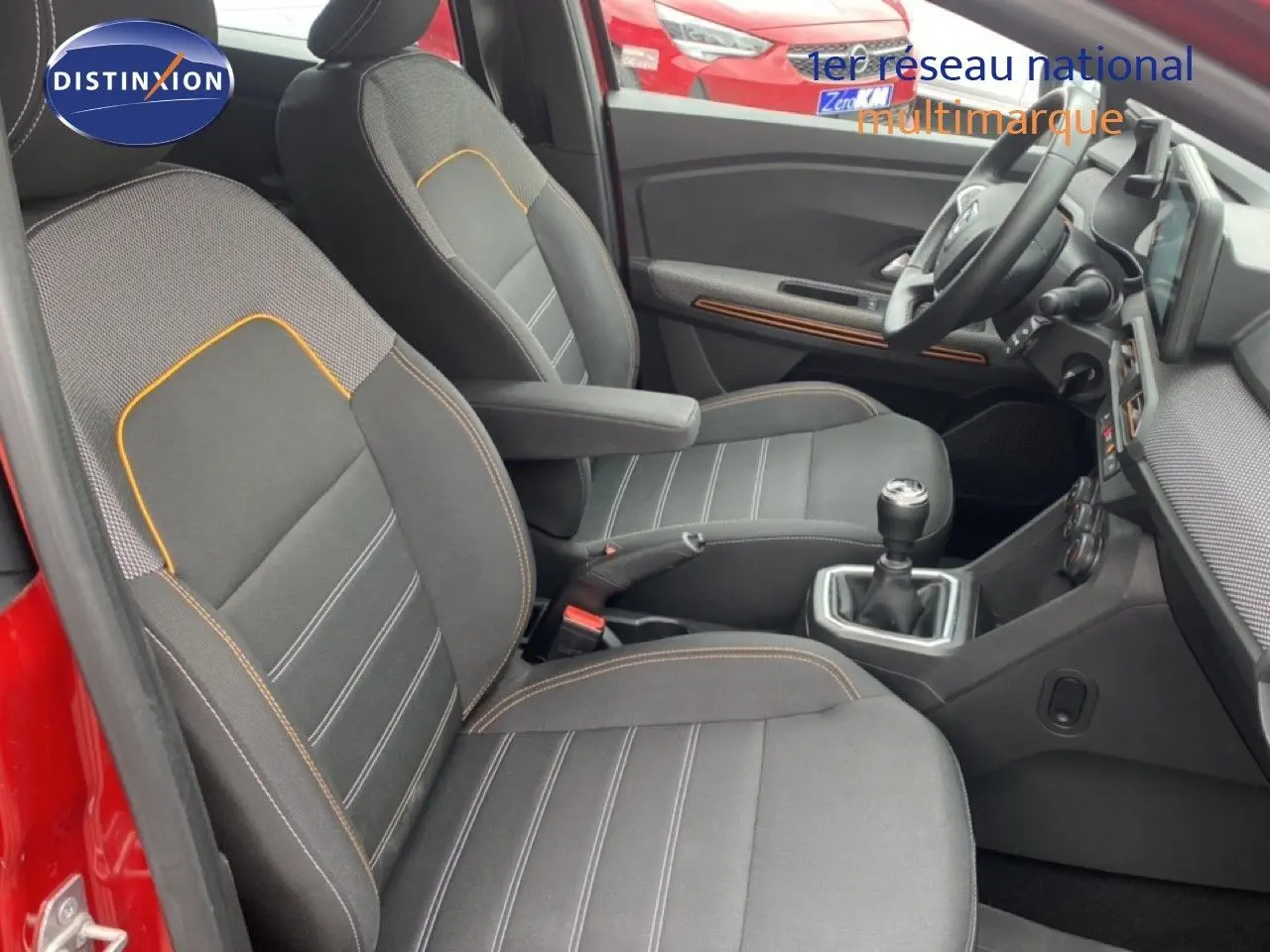 Vue intérieure côté conducteur de la Dacia Sandero rouge fusion, montrant les sièges noirs avec surpiqûres orange et la boîte manuelle.