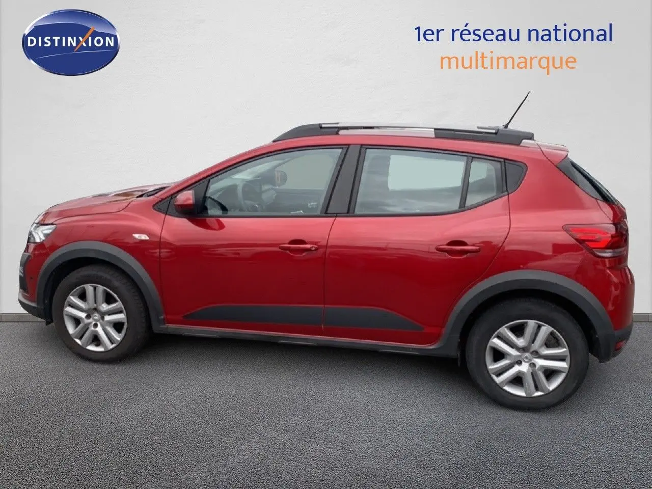 Vue de profil côté gauche d'une Dacia Sandero Stepway rouge fusion métal avec barres de toit et protections noires.