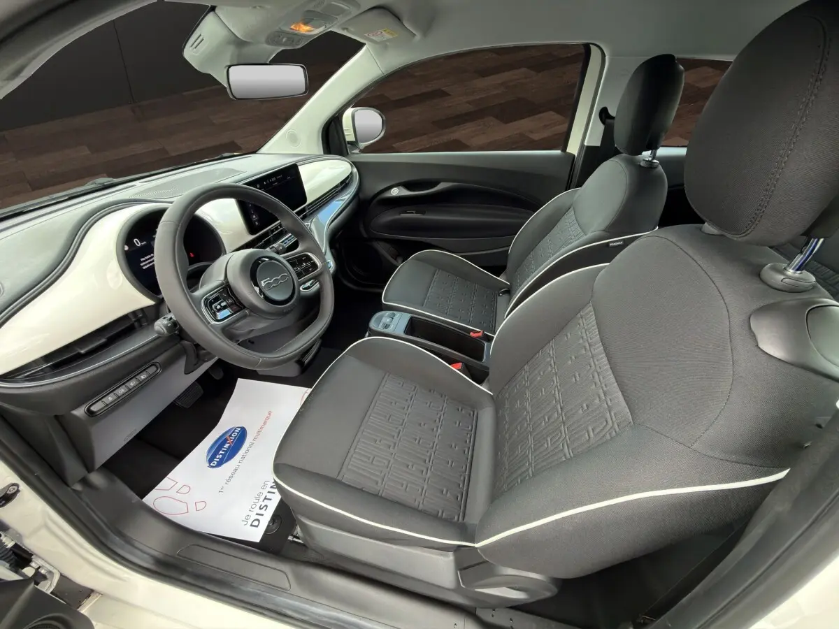Vue intérieure côté conducteur de la Fiat 500 blanche 2023, avec tableau de bord blanc brillant et sièges gris à surpiqûres blanches.