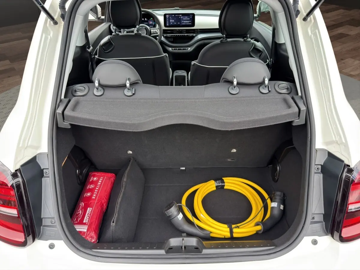 Coffre ouvert d'une FIAT 500 blanche vue de l'arrière, avec câble de charge jaune et trousse de secours rouge visibles.