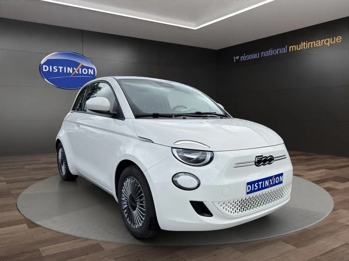 Vue 3/4 avant d'une FIAT 500 électrique blanche avec jantes alu, sur fond intérieur showroom Distinxion.