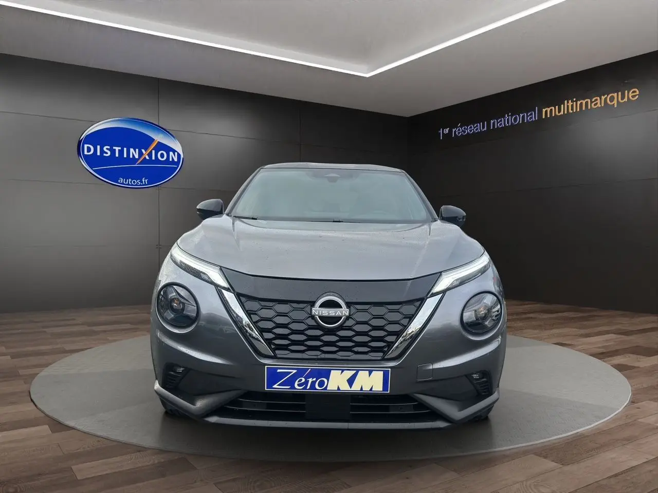 Vue frontale d'un Nissan Juke Full Hybrid gris squale, avec calandre noire et plaque "Zéro KM" en showroom.
