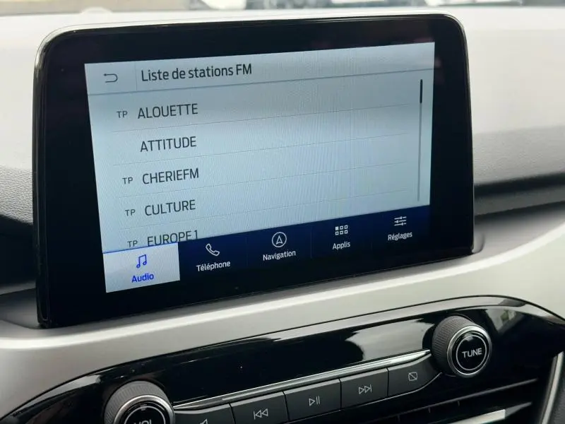 Écran tactile central du Ford Kuga 2024 affichant la liste des stations FM, avec commandes audio et navigation visibles.