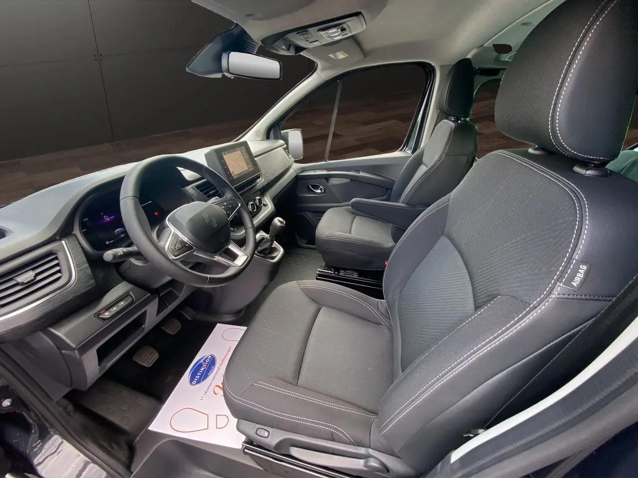 Intérieur avant du Renault Trafic gris comète, vue côté conducteur, sièges noirs avec coutures blanches et tableau de bord moderne.
