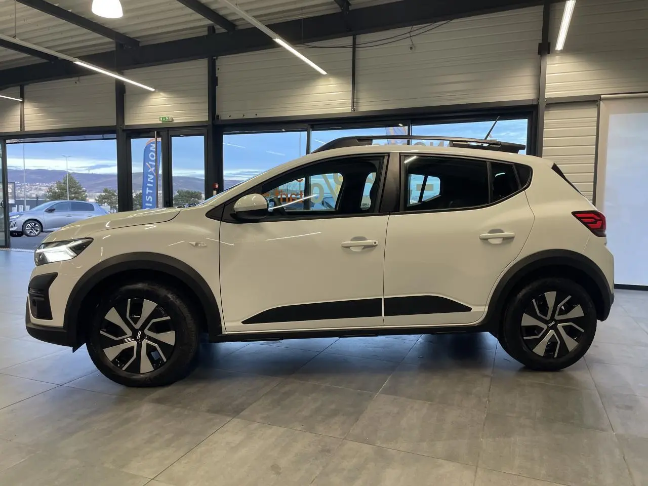 Vue de profil côté gauche d'une Dacia Sandero Stepway blanche Glacier avec jantes noires et barres de toit.