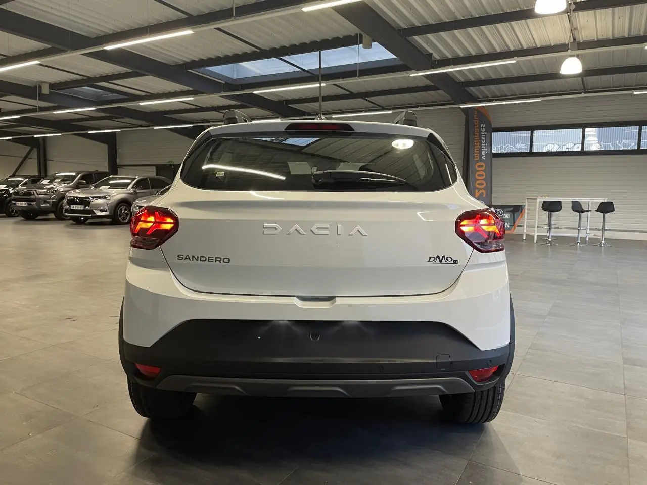 Vue arrière d'une Dacia Sandero TCe 90 Stepway blanc glacier avec feux allumés et bouclier noir en intérieur showroom.