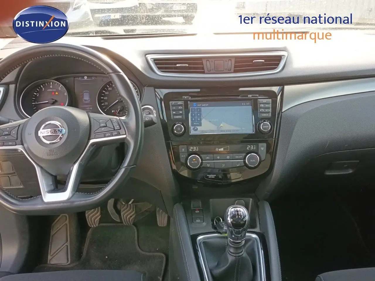 Vue intérieure centrée sur le tableau de bord et la console centrale du Nissan Qashqai blanc, avec volant et levier de vitesse manuel visibles.