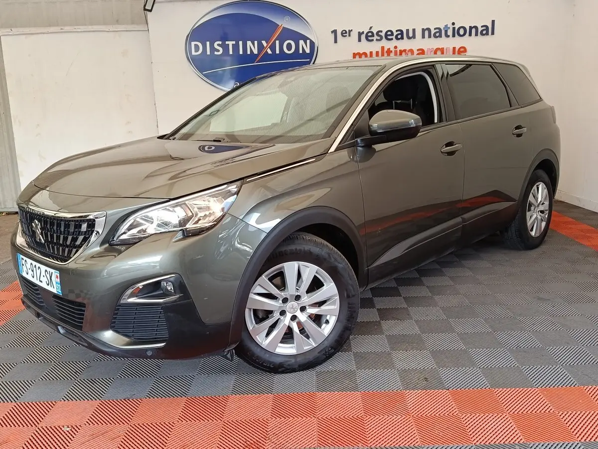 Peugeot 5008 gris foncé vue en 3/4 avant droit, avec phares allumés et jantes alliage, en intérieur showroom.