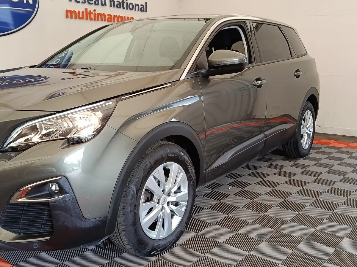 Peugeot 5008 gris foncé vue 3/4 avant droit, avec phares allumés et jantes argentées en intérieur showroom.