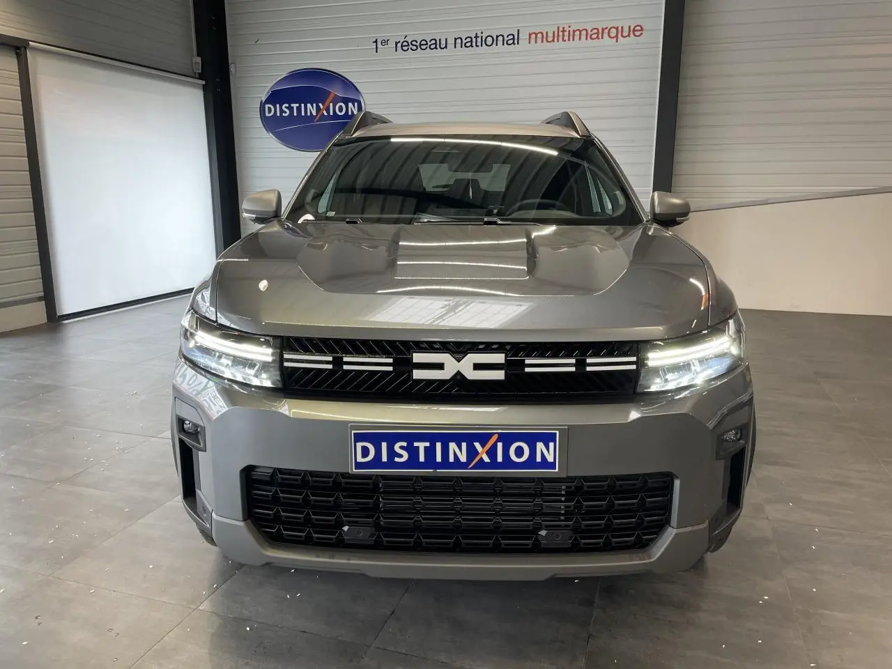 Vue frontale du Dacia Bigster gris schiste avec calandre noire et feux LED allumés en intérieur showroom.