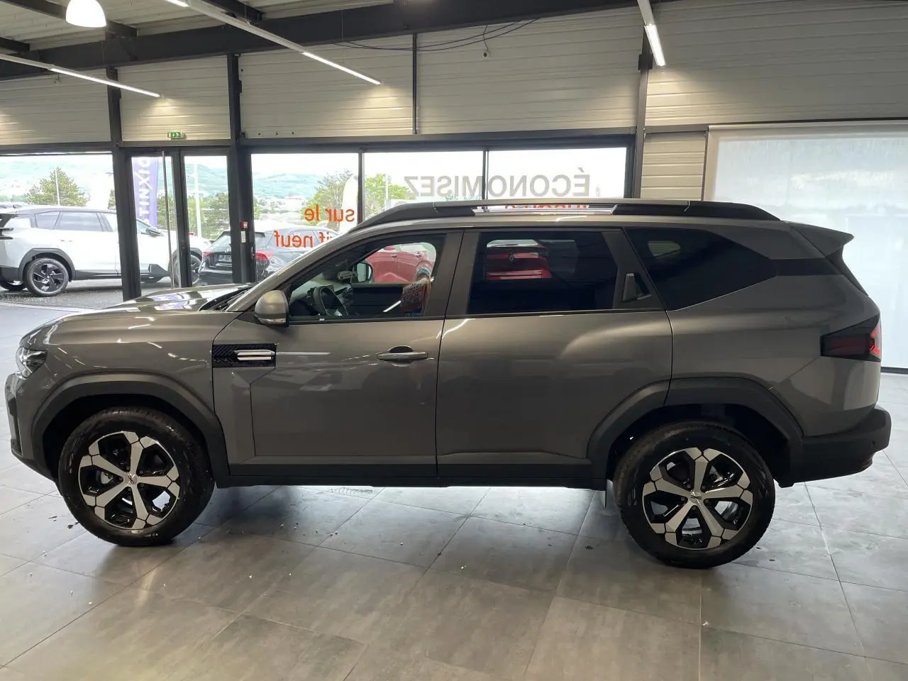 Vue latérale droite du Dacia Bigster gris schiste 2026 avec jantes alliage 18 pouces et barres de toit noires.