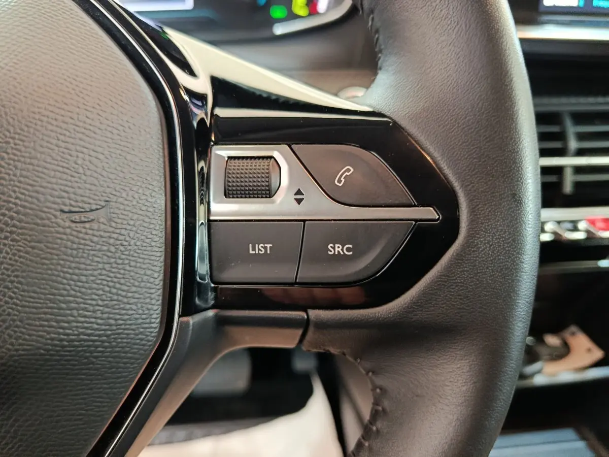 Gros plan sur les commandes gauche du volant cuir noir du Peugeot 208 électrique blanc, avec boutons multifonctions.