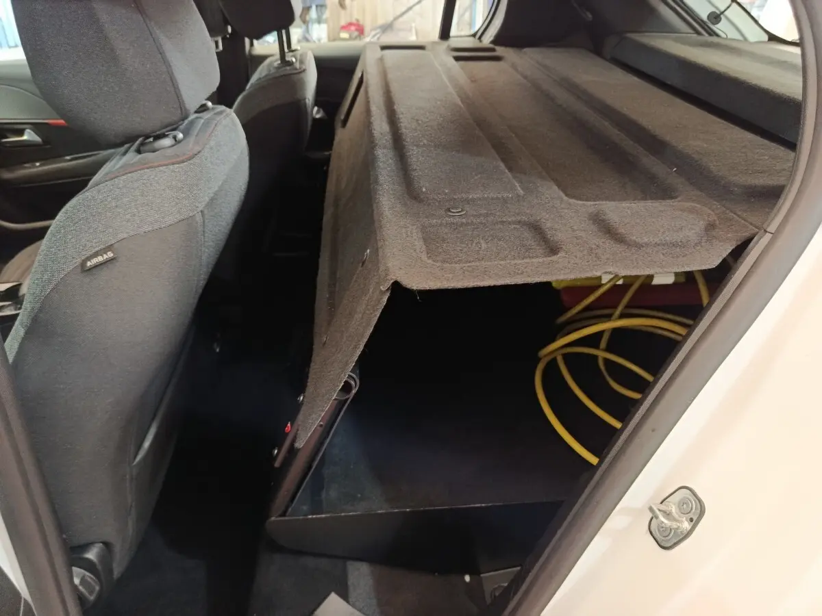 Vue intérieure côté droit arrière du coffre ouvert d'une Peugeot 208 électrique blanche, avec câble de recharge visible.