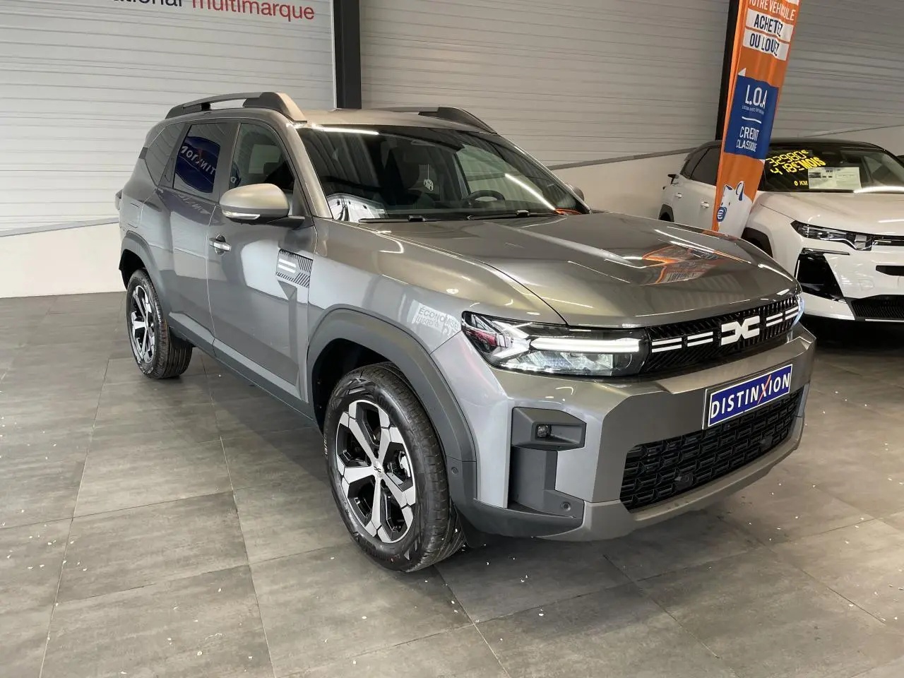 SUV Dacia Bigster gris schiste en 3/4 avant droit, avec jantes alliage et barres de toit visibles en intérieur showroom.