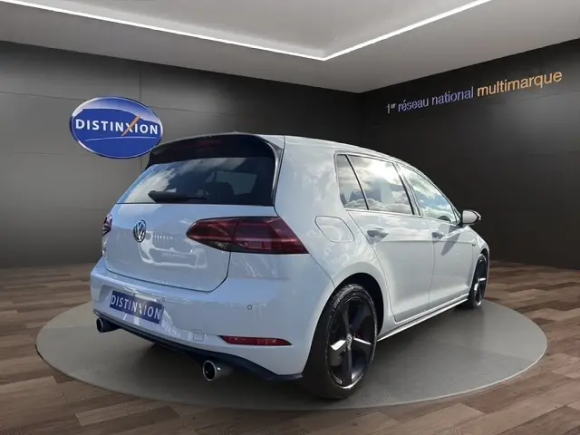 Vue de profil droit d’une Volkswagen Golf GTI Performance blanche avec jantes noires et étriers rouges, en intérieur showroom.