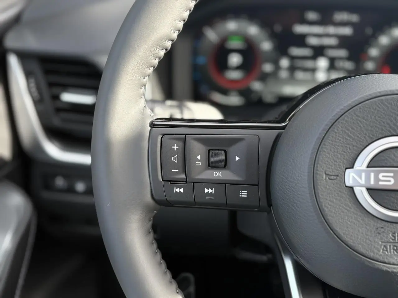 Gros plan sur les commandes au volant à gauche du Nissan Qashqai blanc nacré, avec tableau de bord numérique flou en arrière-plan.