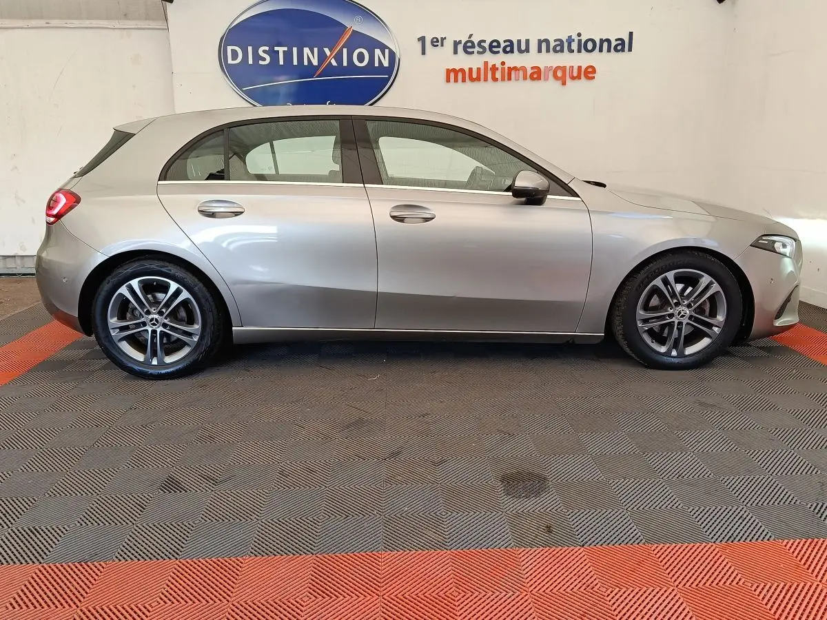 Vue de profil côté gauche d'une Mercedes Classe A 180 Business Line gris clair stationnée en intérieur sur sol à damiers.
