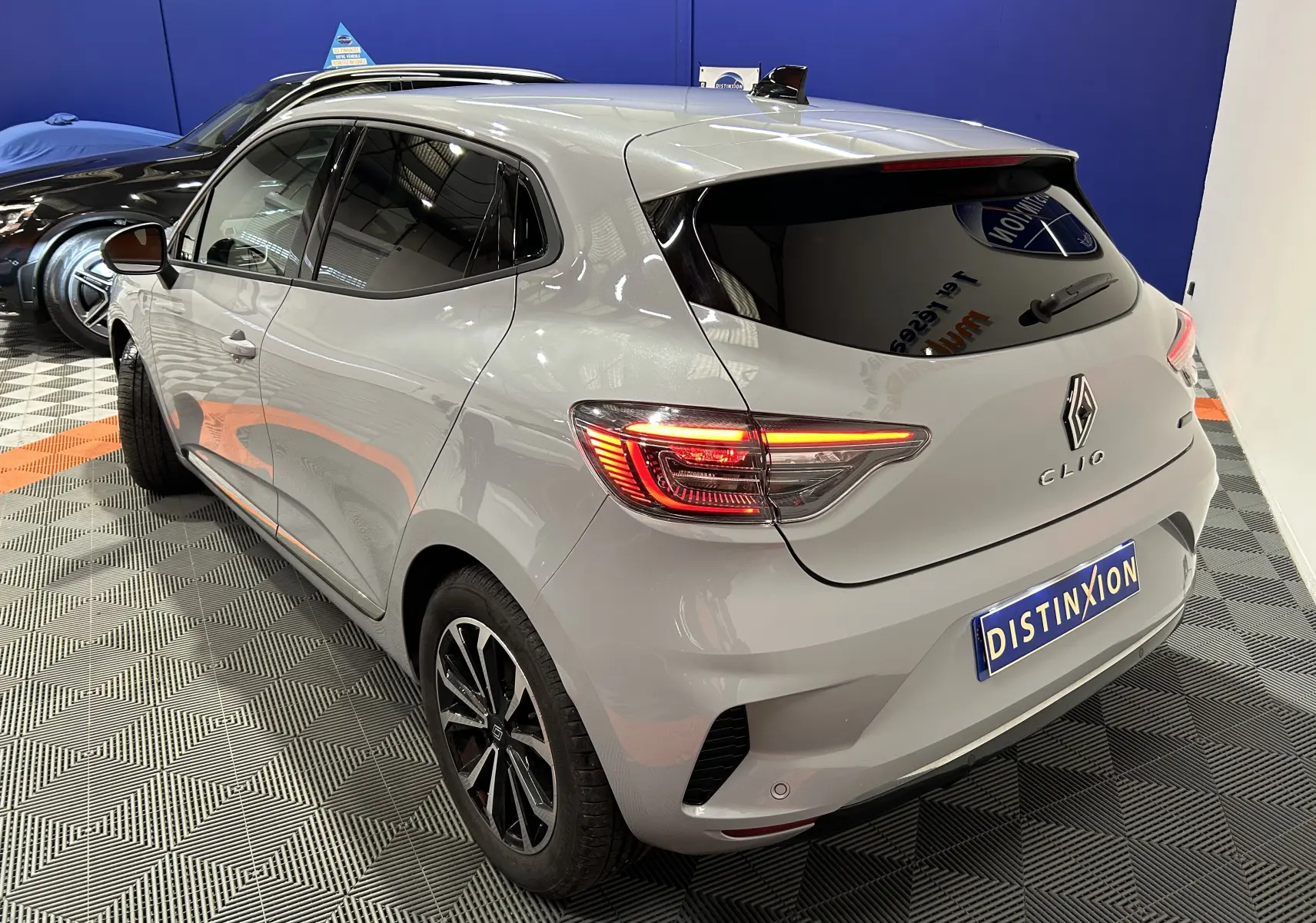 Vue 3/4 arrière d'une Renault Clio V E-Tech gris Rafale métallisé avec feux arrière allumés dans un showroom.