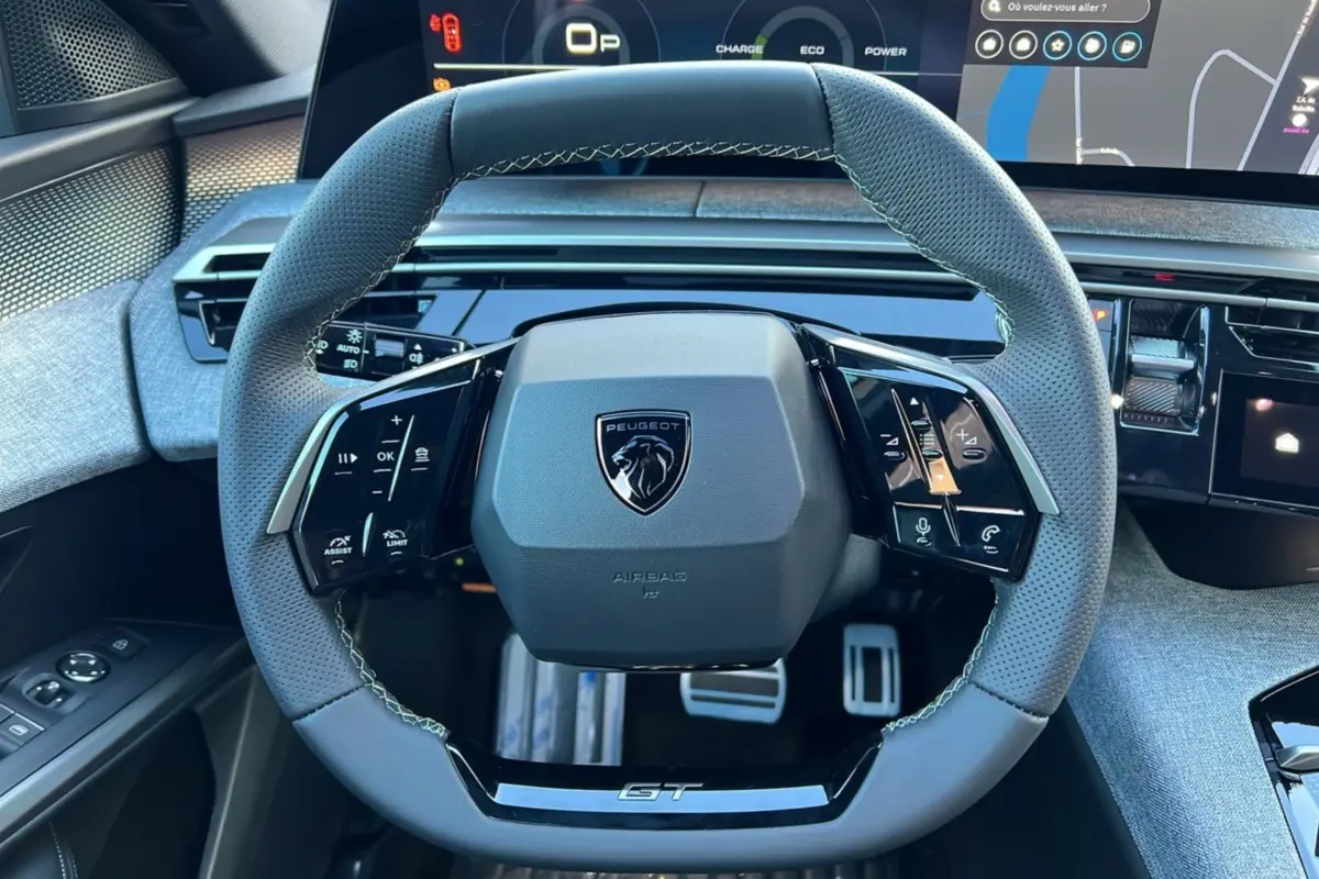 Vue rapprochée du volant cuir perforé gris avec logo Peugeot et inscription GT, tableau de bord numérique en arrière-plan.