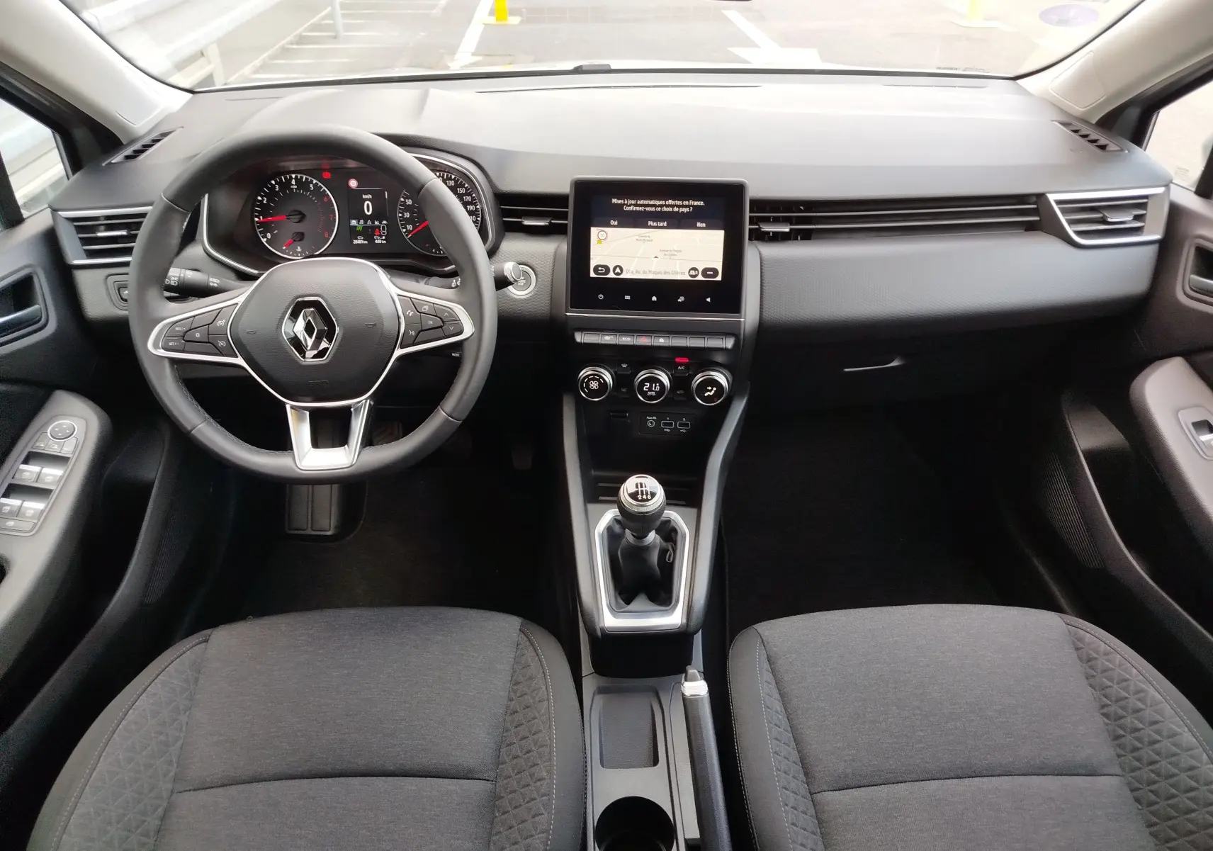 Vue intérieure avant centrée sur le tableau de bord et volant de la Renault Clio V Business, avec écran tactile et levier de vitesses manuel.