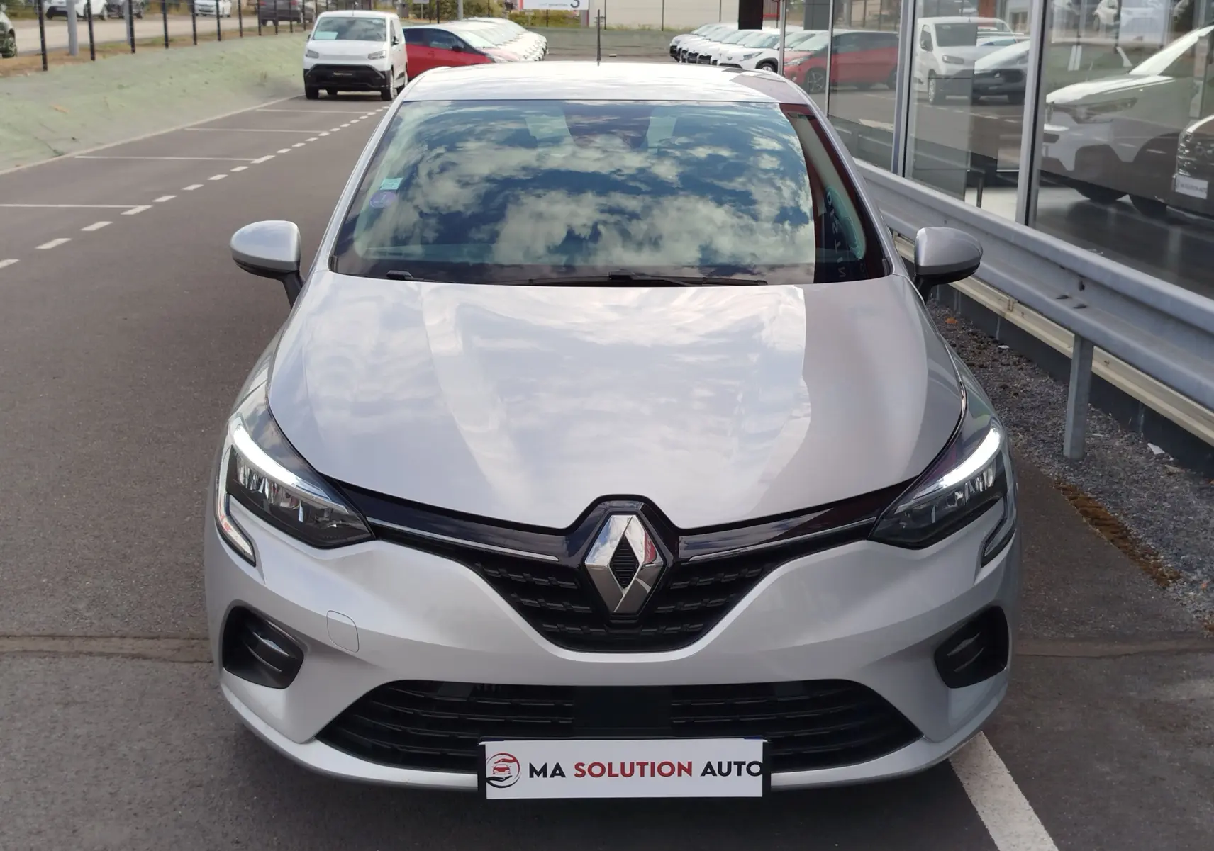 Vue avant d'une Renault Clio V gris platine, avec calandre noire et phares LED allumés, stationnée devant un bâtiment vitré.