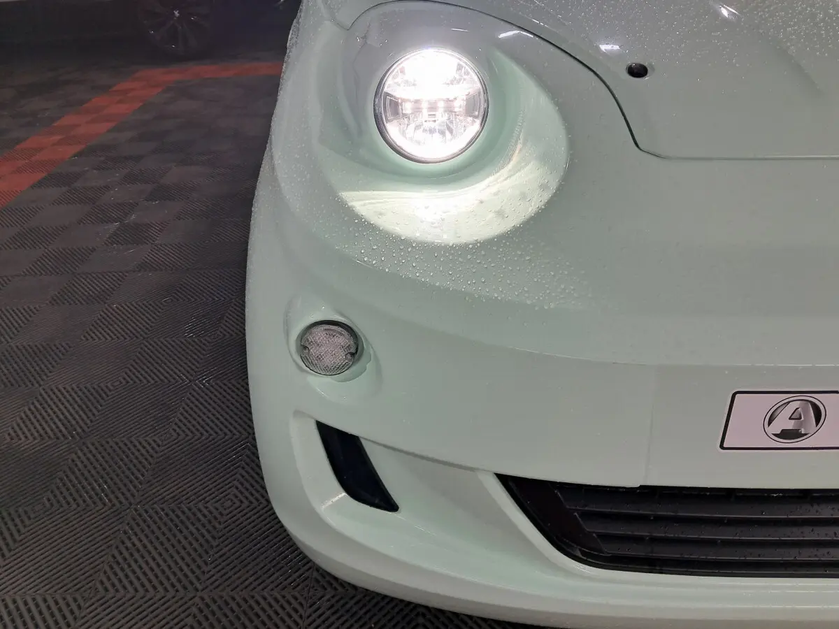 Gros plan sur l'avant gauche vert d'une Aixam MINAUTO E chic 2025 avec phare allumé et logo visible.