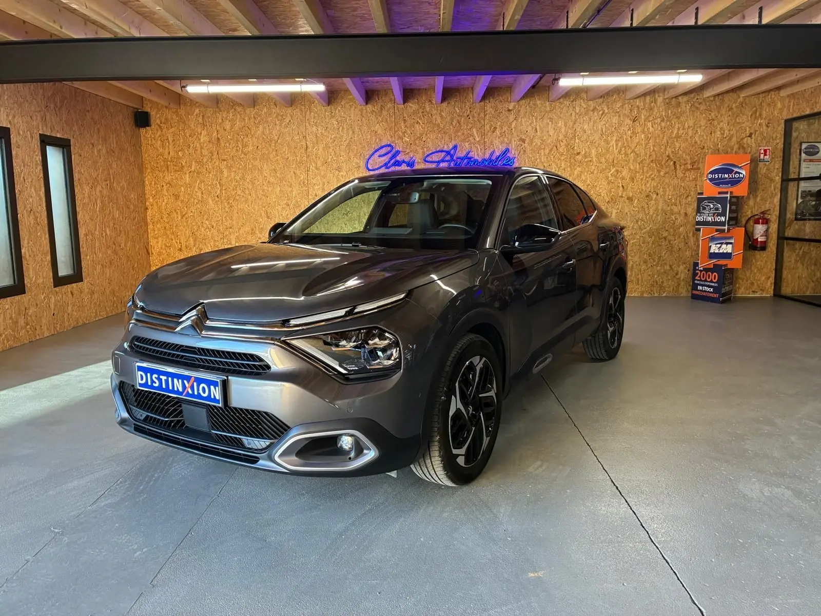 Vue 3/4 avant droite d'une Citroën C4 X gris métallisé 2024 avec phares LED et jantes noires en intérieur showroom.