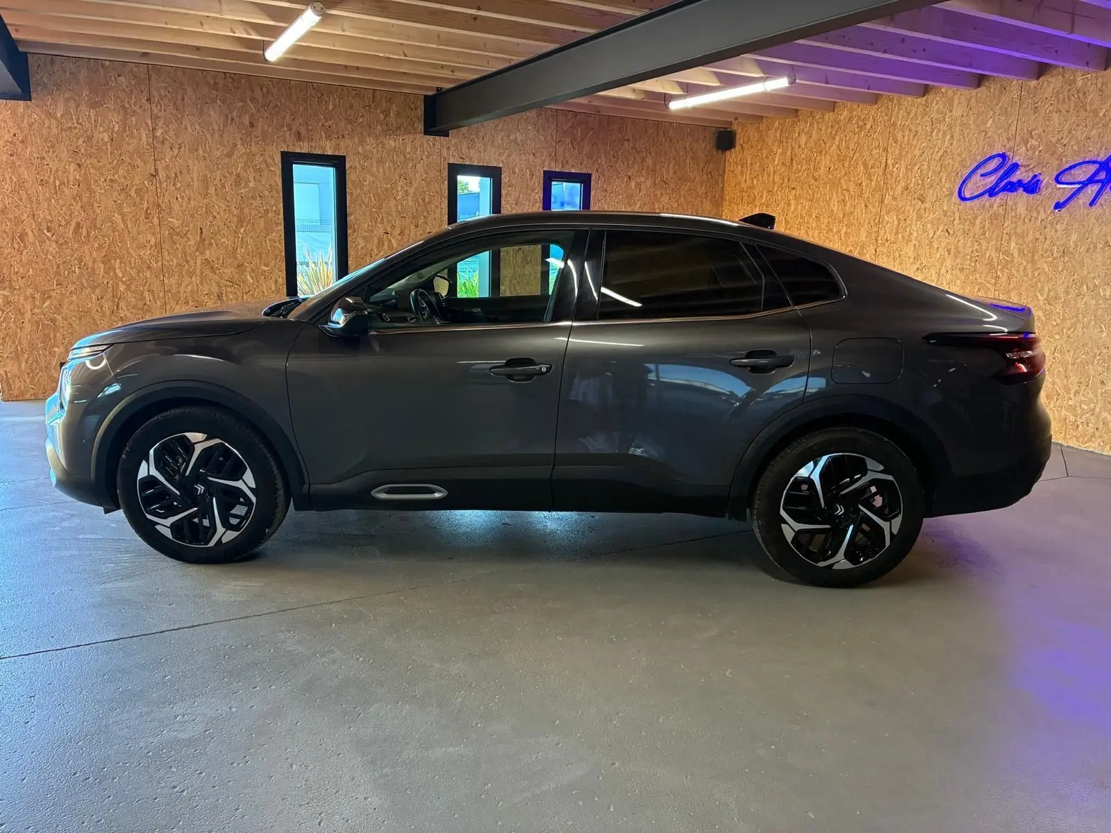 Vue de profil côté gauche d'une Citroën C4 X gris foncé 2024 avec jantes noires et vitres teintées dans un intérieur en bois.