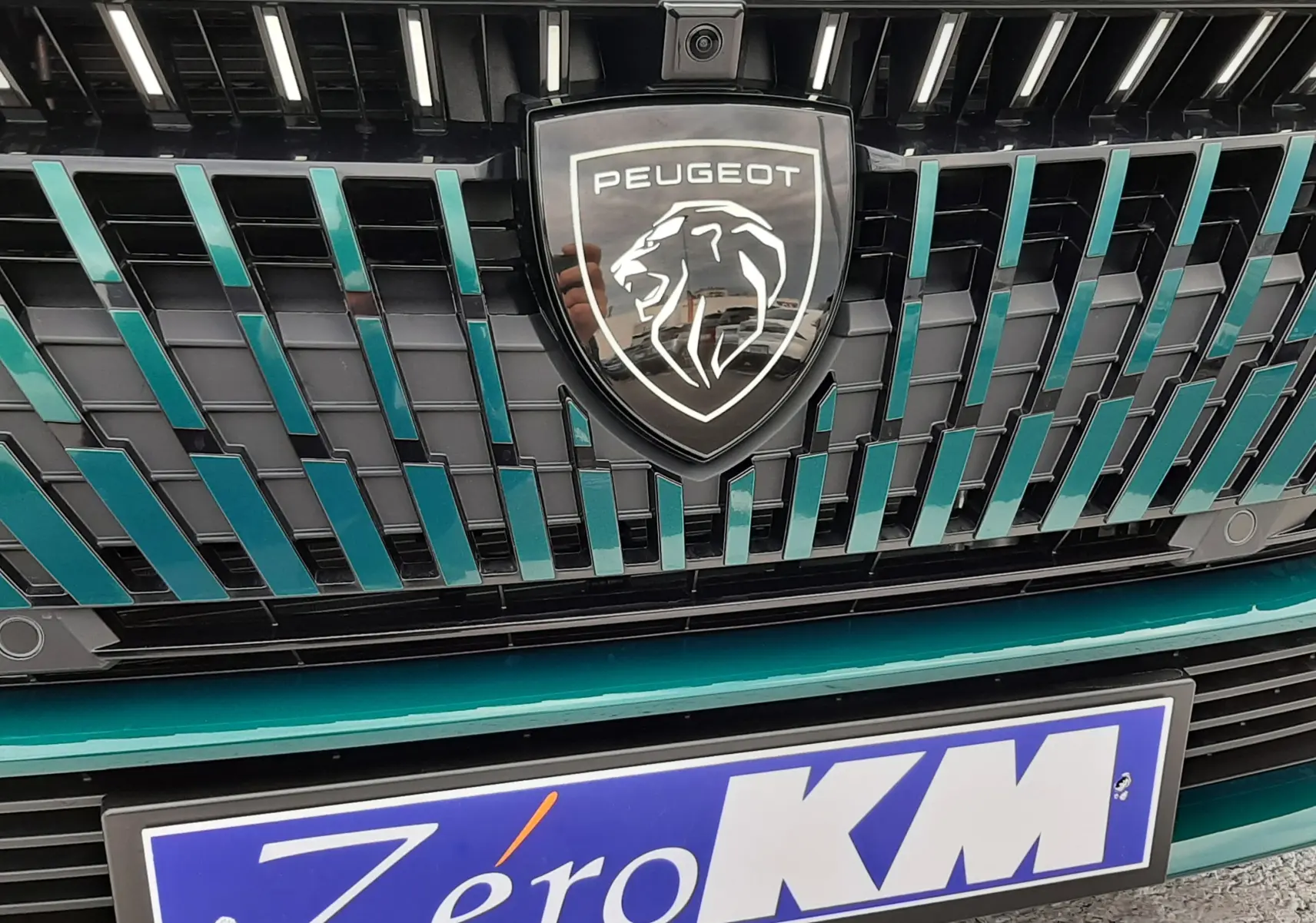 Gros plan sur la calandre bleu Lagoa d'une Peugeot 308 2025 avec logo lion et plaque Zéro KM visible.
