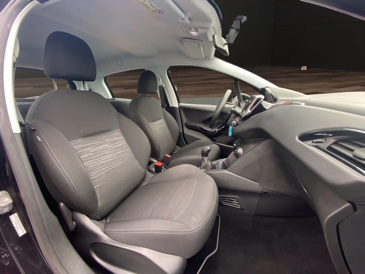 Vue intérieure côté conducteur de la Peugeot 208 Affaire noire, sièges tissu noir et tableau de bord moderne.