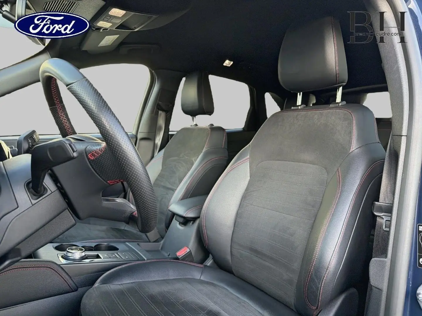 Vue intérieure côté conducteur du Ford Kuga 2023 bleu, sièges sport noirs avec surpiqûres rouges et volant cuir perforé.