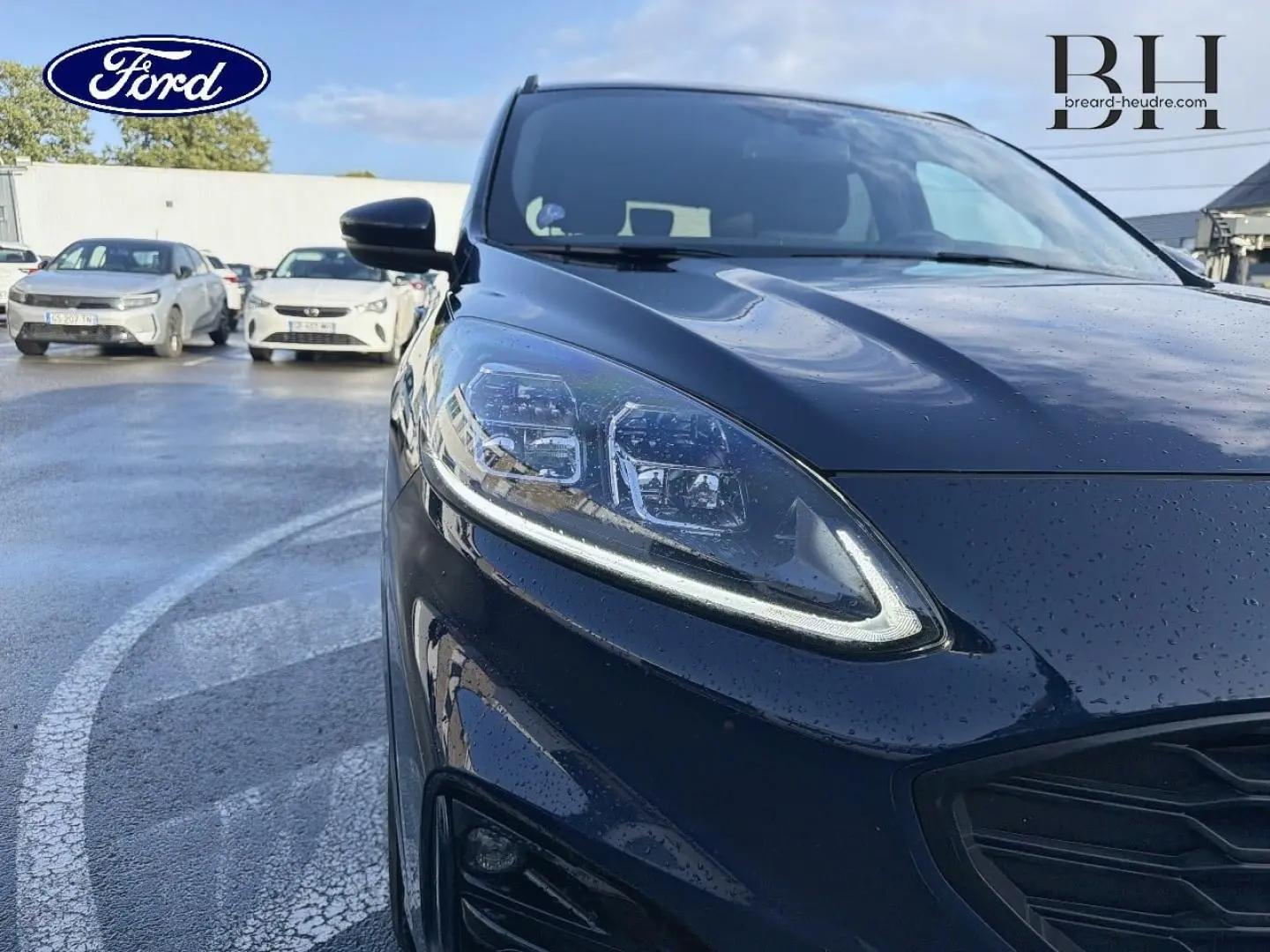 Gros plan sur l'avant gauche du Ford Kuga bleu avec phares LED et calandre noire brillante sous la pluie.