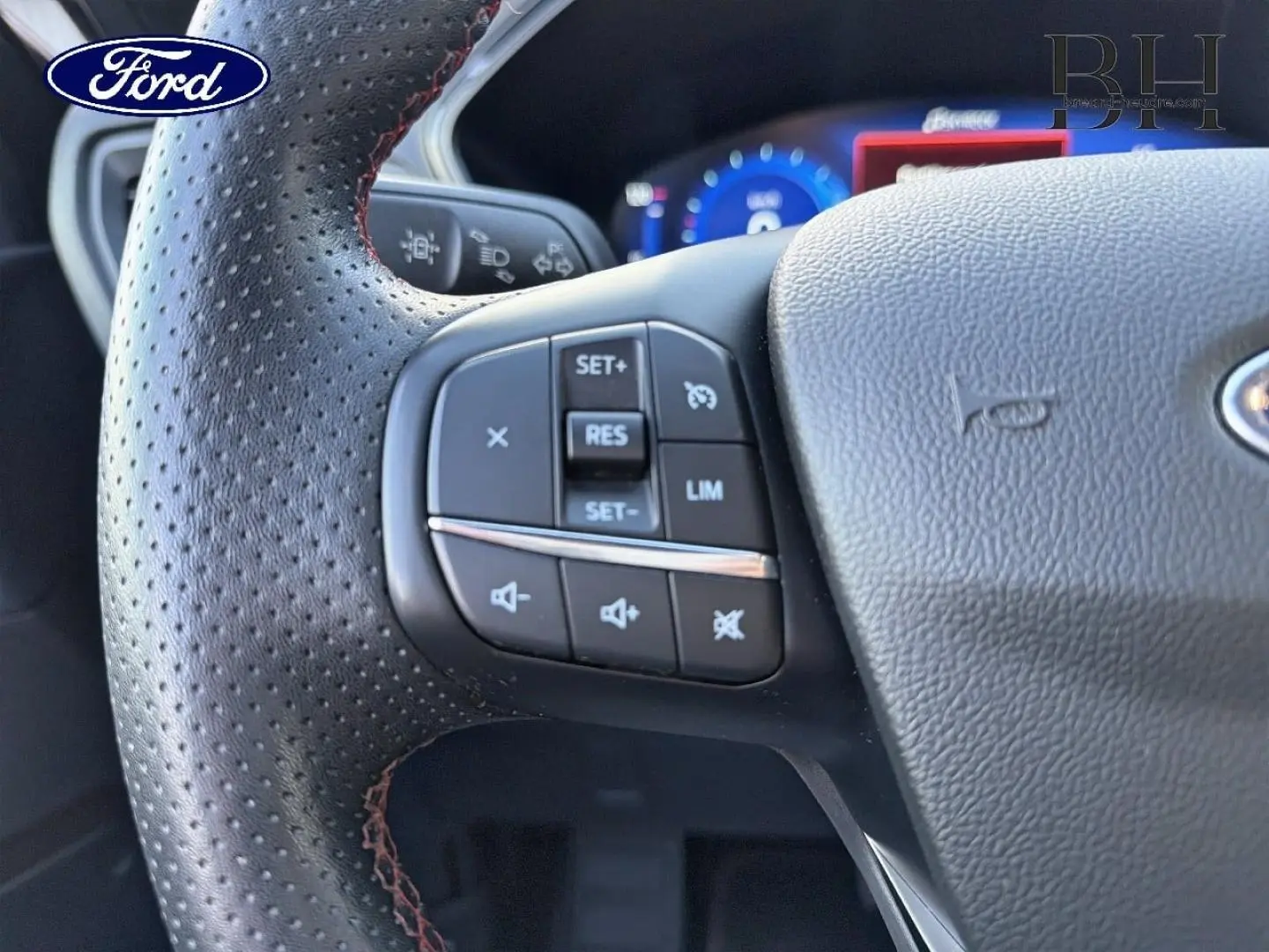 Gros plan sur le volant cuir perforé du Ford Kuga 2023 avec commandes multifonctions côté gauche et tableau de bord numérique bleu.