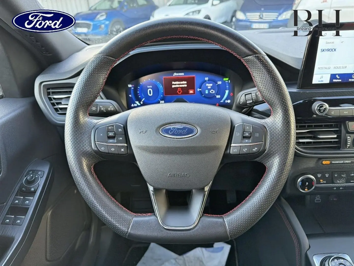 Vue centrée sur le volant cuir perforé avec surpiqûres rouges du Ford Kuga bleu, tableau de bord numérique et écran tactile visible.