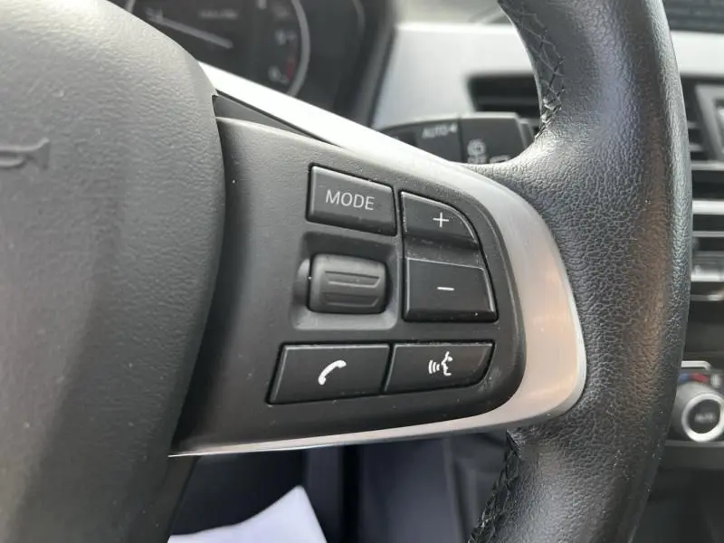 Gros plan sur les commandes multifonctions à gauche du volant noir du BMW X1 20D 2017, avec boutons mode, volume et téléphone.