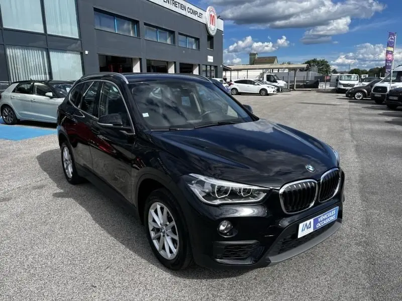 BMW X1 20D noir métal en 3/4 avant droit, avec calandre chromée et jantes alliage visibles sur parking extérieur.