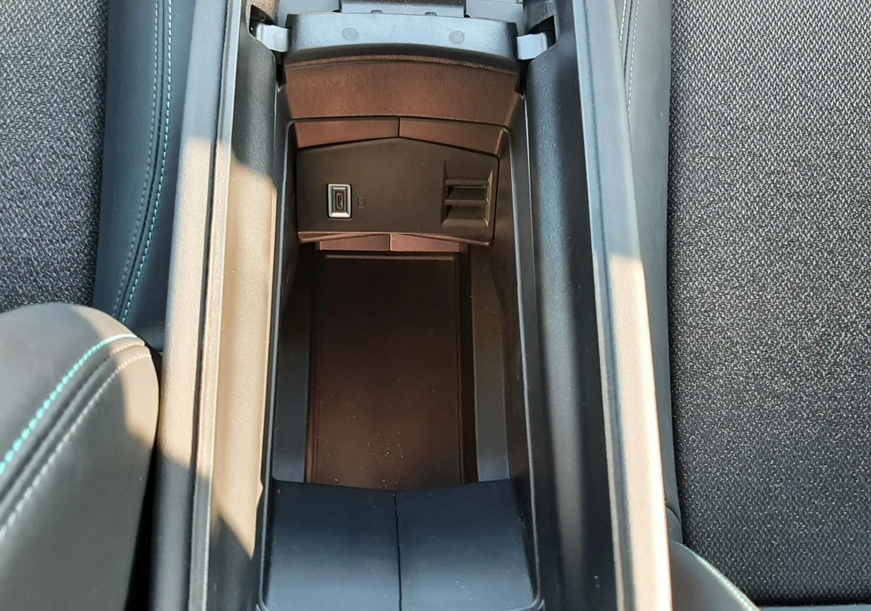 Vue plongeante sur le rangement central ouvert de la Peugeot 308 SW Hybrid 2025, avec prise USB visible et sellerie noire.
