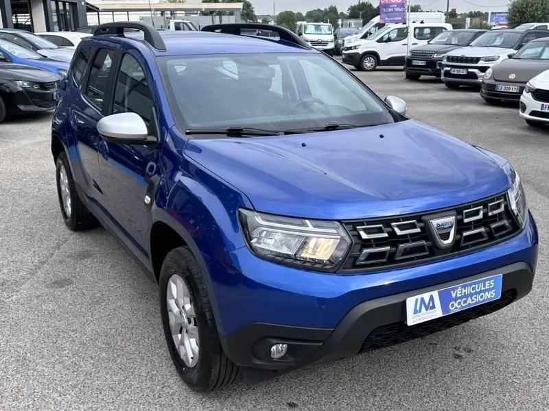 Vue 3/4 avant gauche d'un Dacia Duster bleu métal 2022 avec calandre noire et rétroviseurs argentés sur parking.