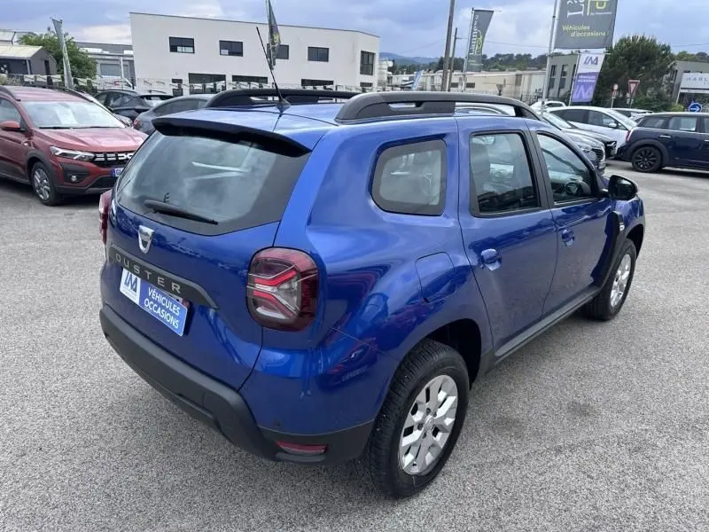 Vue 3/4 arrière droite d'un Dacia Duster 2022 bleu métal, version ECO-G 100 ch, avec barres de toit et feux arrière distinctifs.
