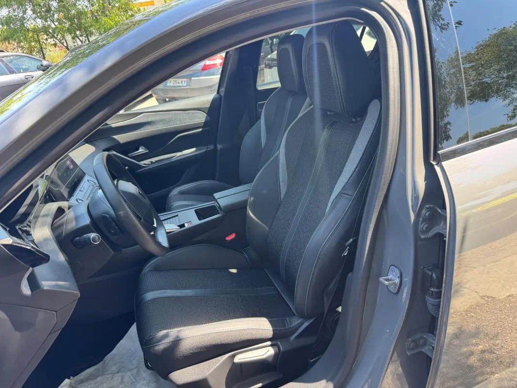 Vue intérieure côté conducteur d'une Peugeot 308 gris 2025, sièges noirs avec accoudoir central et volant multifonction.